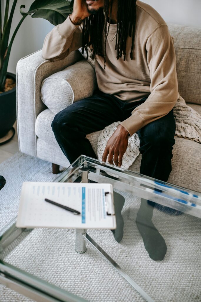 Unrecognizable African American male with dreadlocks leaning on hand while sitting on couch near table during psychotherapy session in office